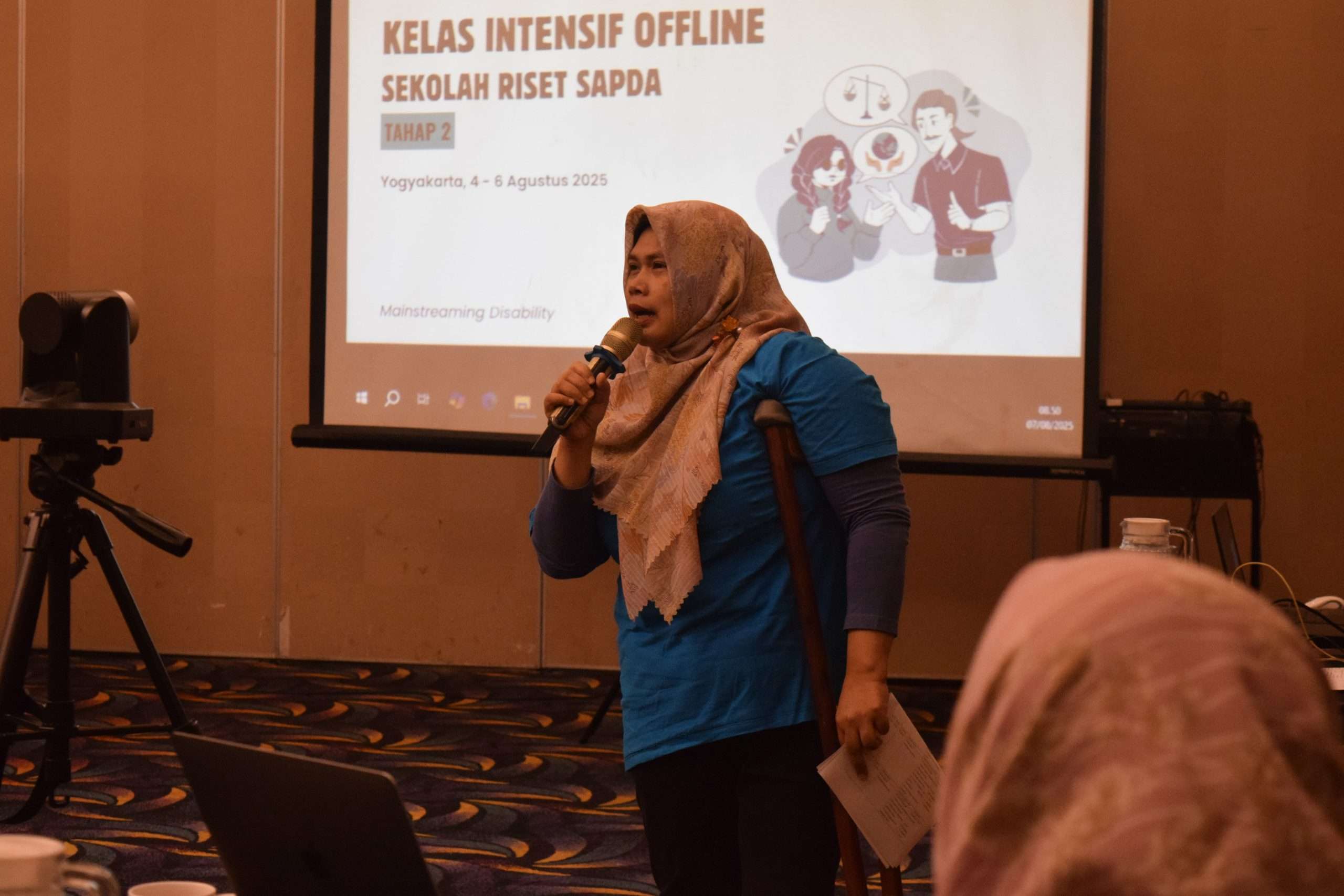 Nurul Saadah, Direktur SAPDA, mengenakan baju berwarna biru dan membawa mic. Nurul sedang berbicara dalam rangka menyambut peserta kelas riset offline