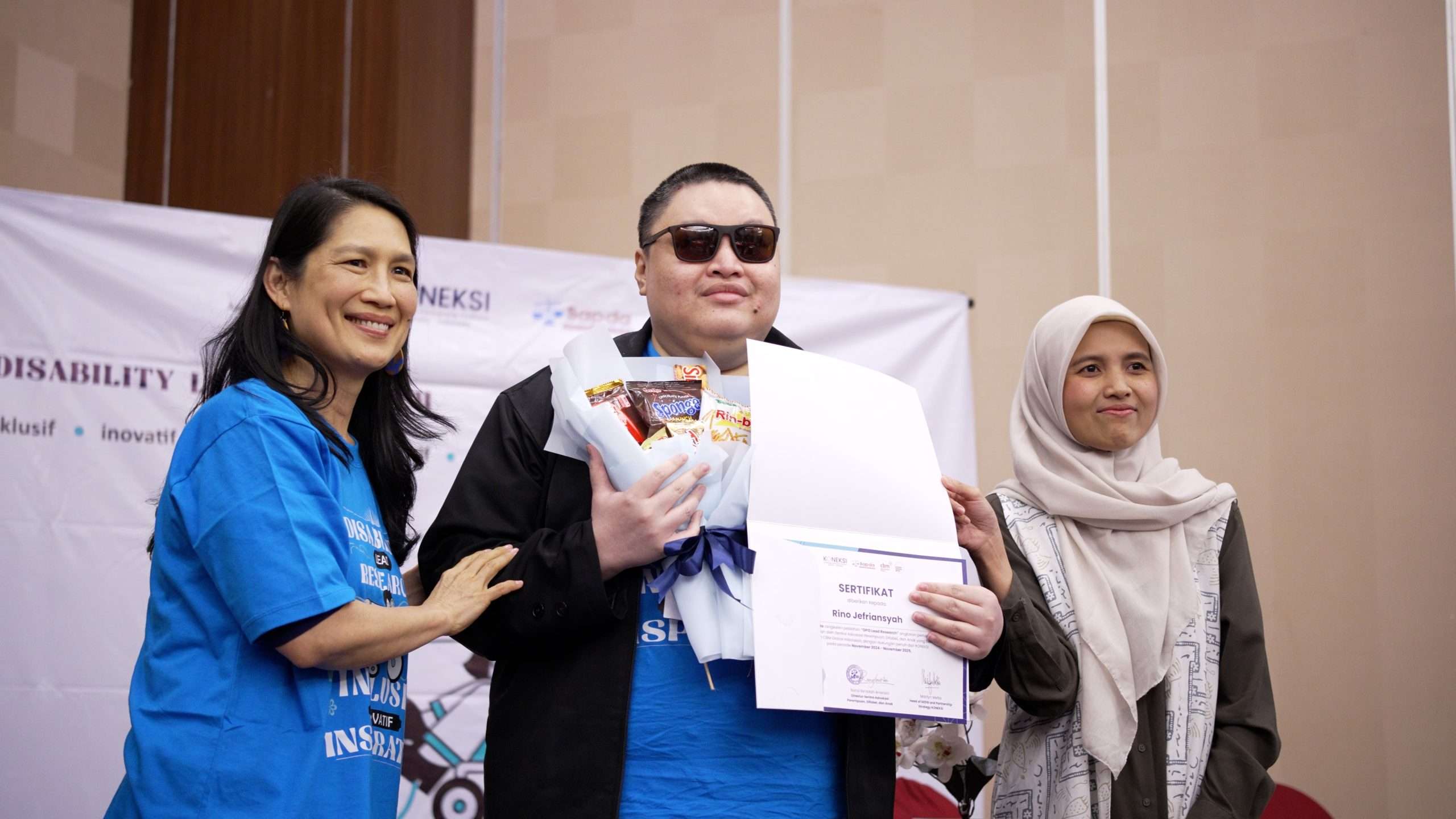 Laki laki penyandang disabilitas netra memegang sertifikat kelas riset penyandang disabilitas dengan ditemani oleh dua perempuan dari perwakilan KONEKSI dan Bappenas dalam acara Sekolah Riset SAPDA